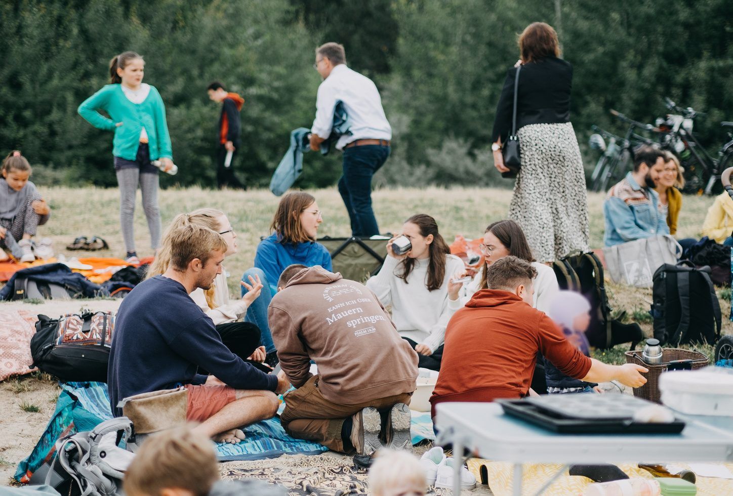 A group of young people sitting on blankets in a meadow, chatting, with trees and bicycles in the background