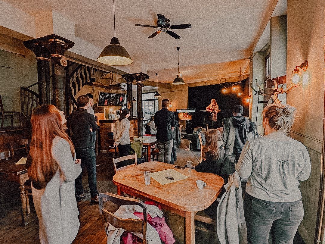 People standing in a café with wooden tables and pendant lamps, listening to a singer with a guitar on a small stage