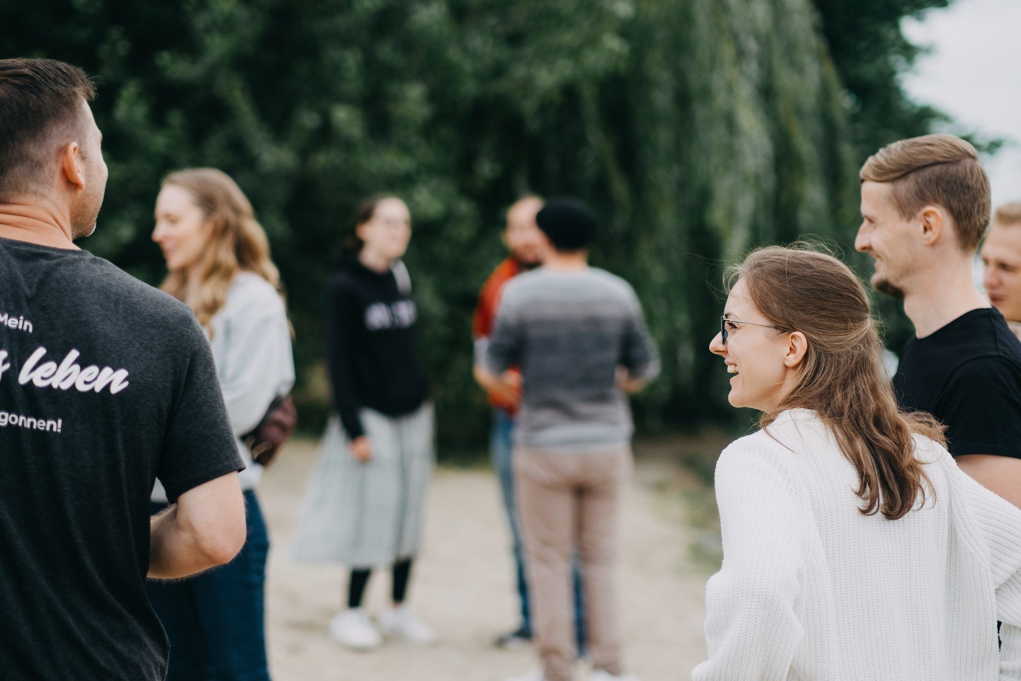 A young woman with glasses laughs while talking with a group of young adults standing outdoors in front of trees