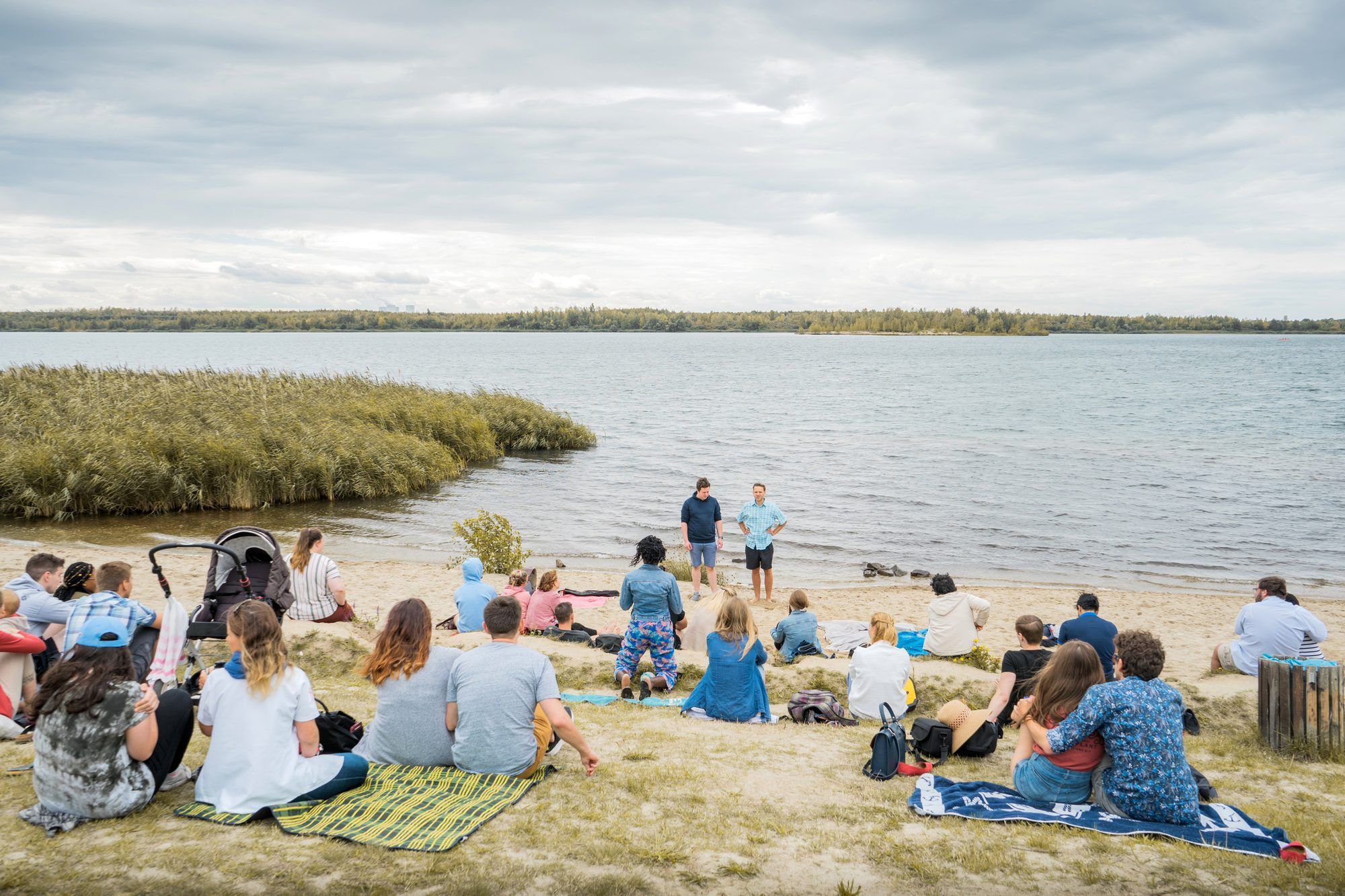 A group sitting on blankets on a sandy lakeshore, listening to two men standing at the water’s edge