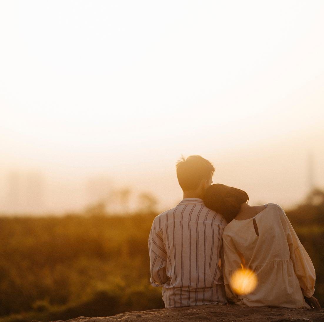 A man and a woman seen from behind sitting side by side in warm evening light, her head resting on his shoulder