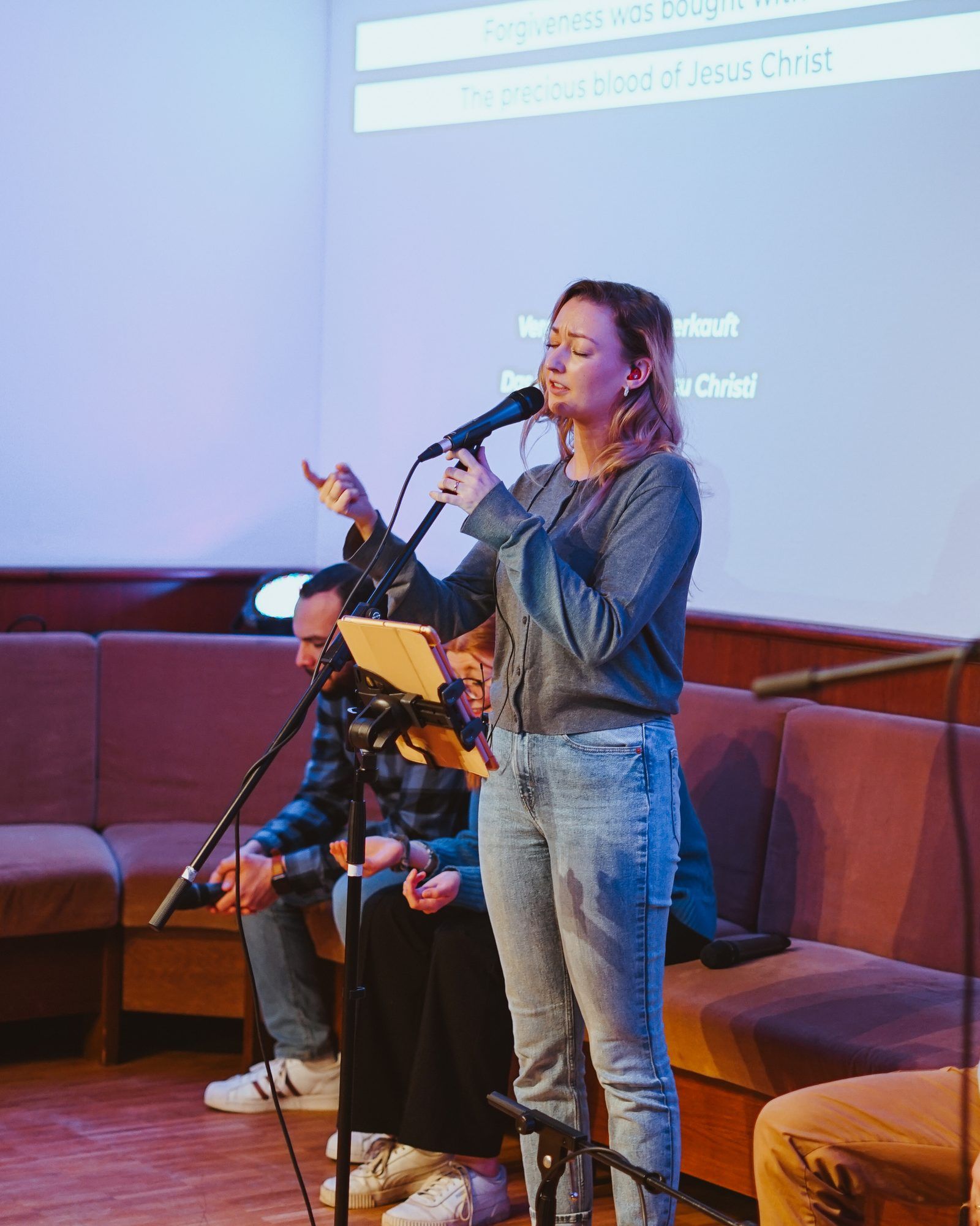 A woman singing into a microphone with her eyes closed, people sitting on a bench behind her in front of a screen showing song lyrics