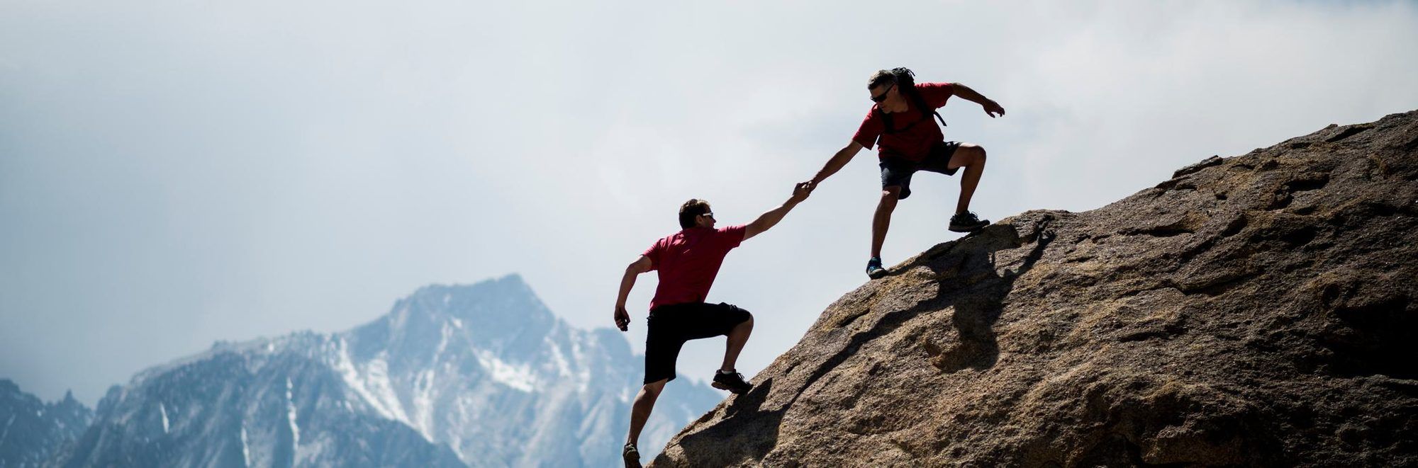A climber reaching down to pull another up a steep rock face, with snow-capped mountains in the background