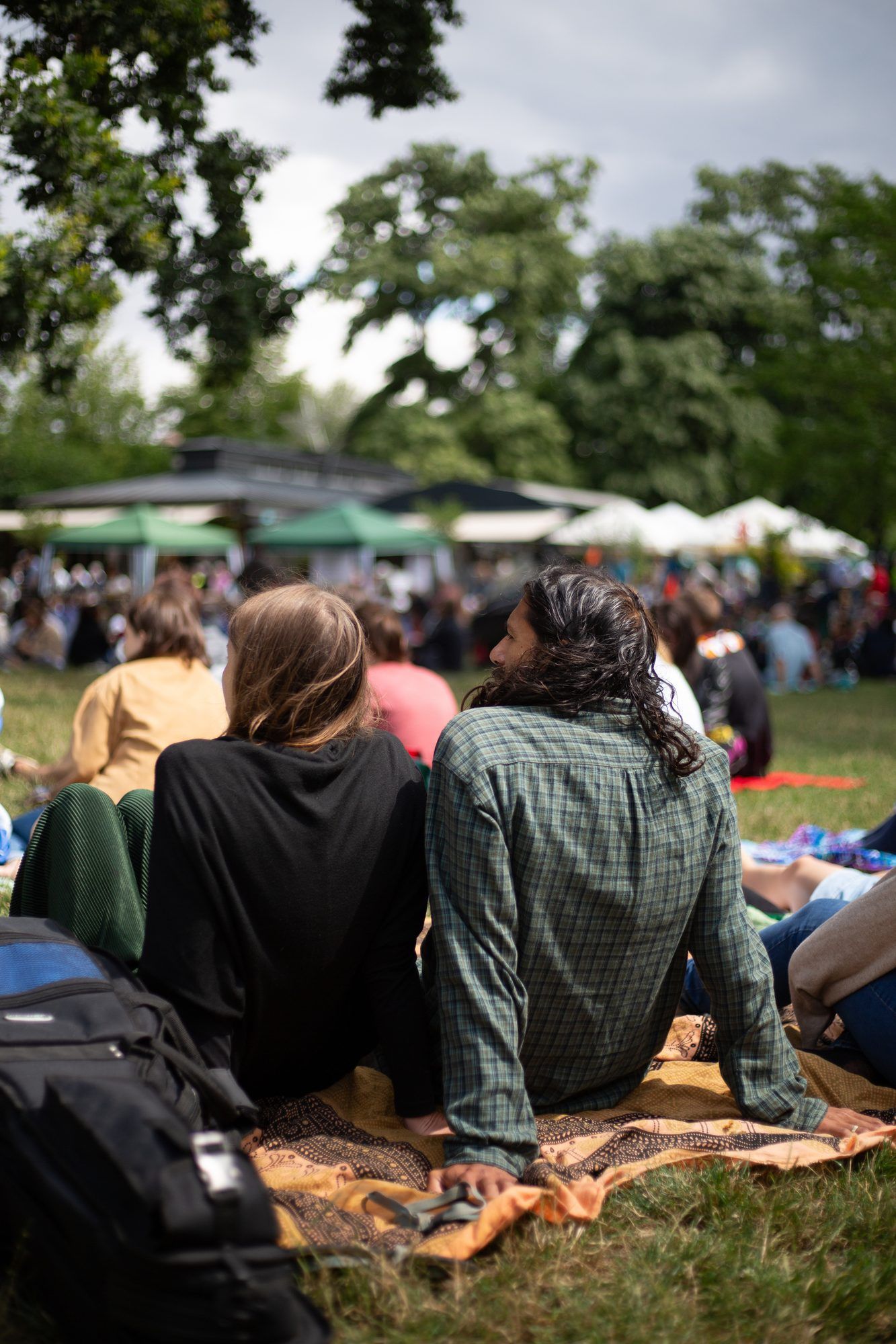 Two people seen from behind sitting on a blanket on the grass, with many more people and tents ahead of them in a park