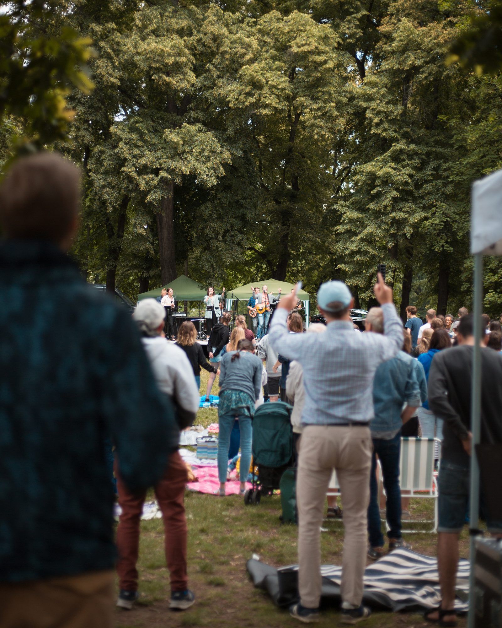 People at an open-air church service in a park, a man in front raising his hands, with a band playing under green gazebos in the background