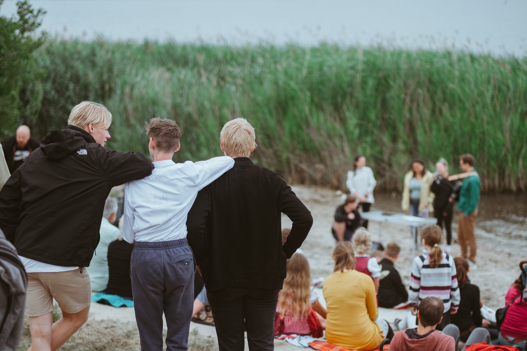 Three young men stand arm in arm on a shore in front of tall reeds, watching a group sitting on the sand