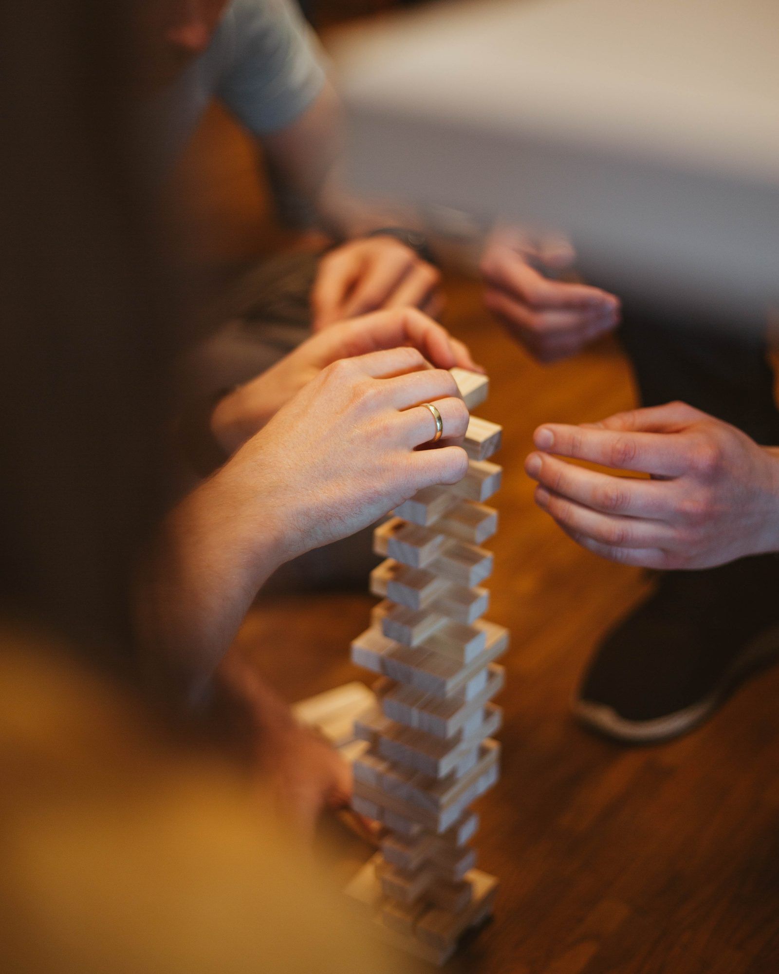 Several hands working together on a tall tower of wooden blocks on a wooden table