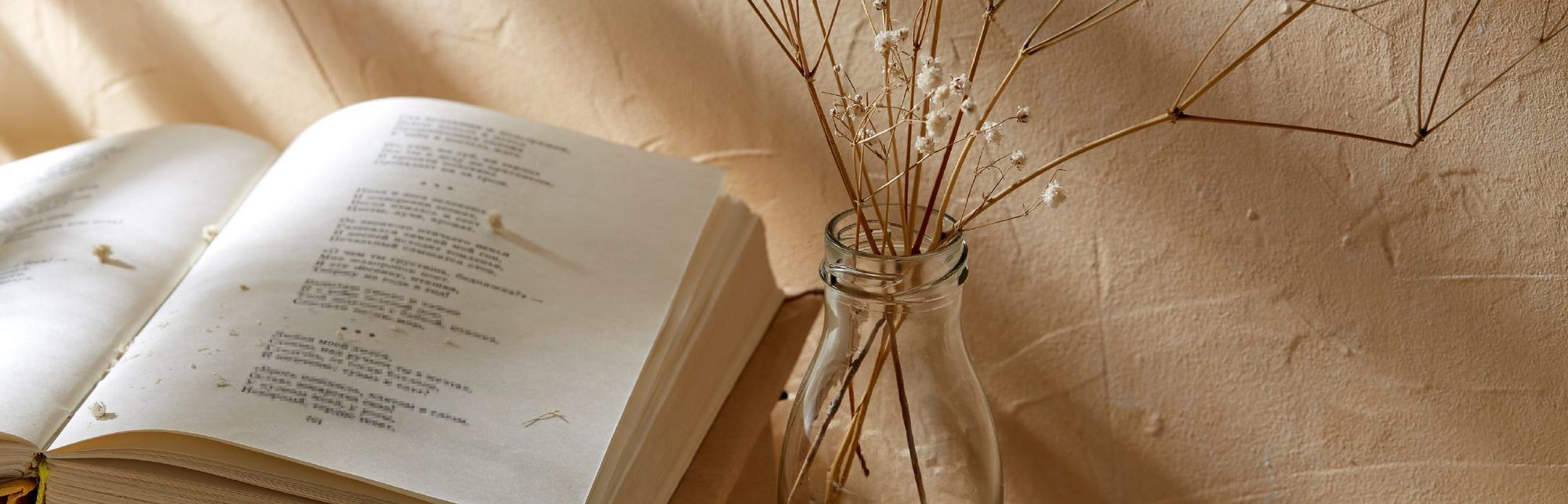 Open book of poems next to a glass bottle with dried flowers in front of a beige wall