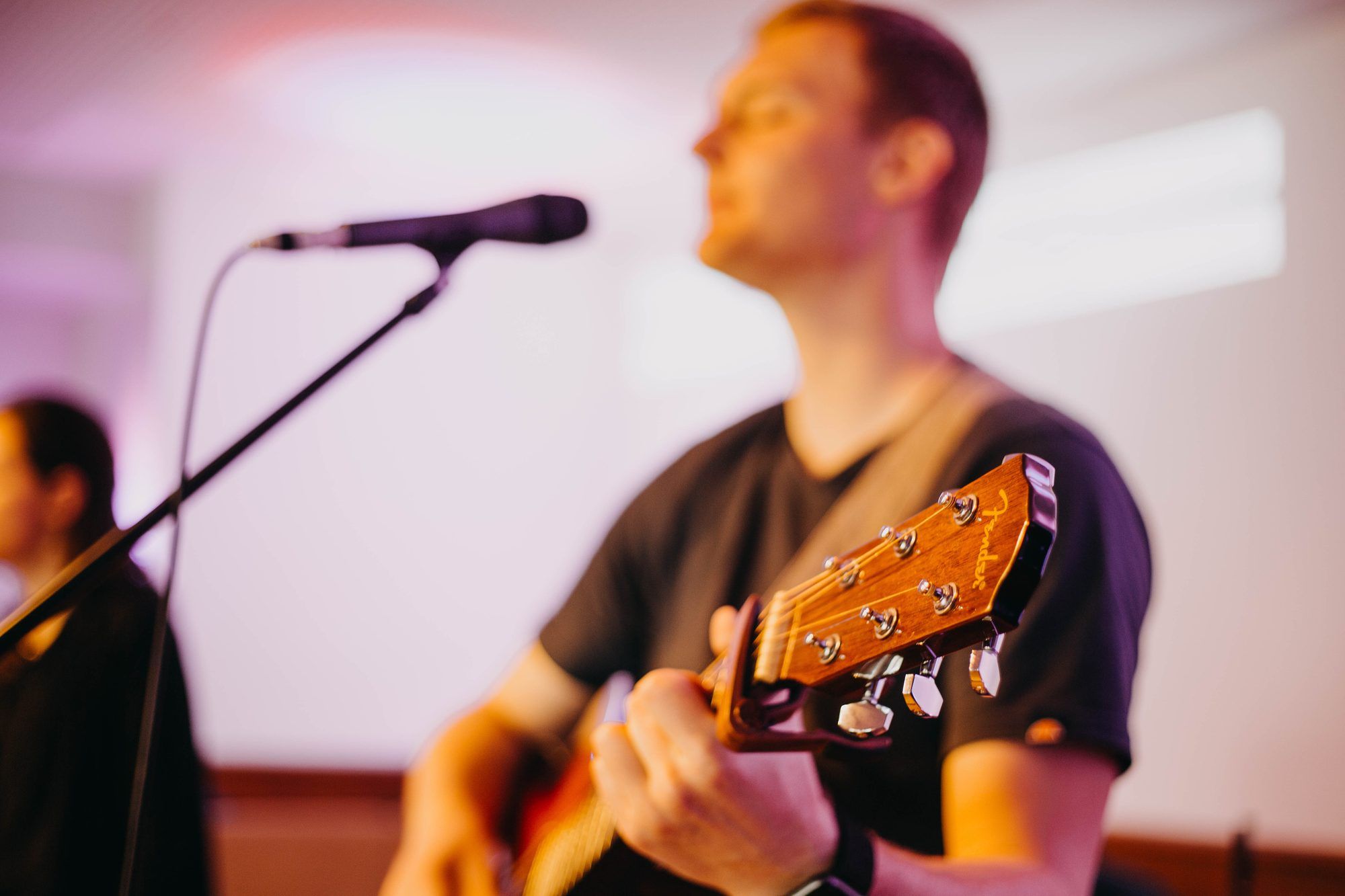 A man playing acoustic guitar and singing at a microphone, with the guitar headstock in sharp focus in the foreground
