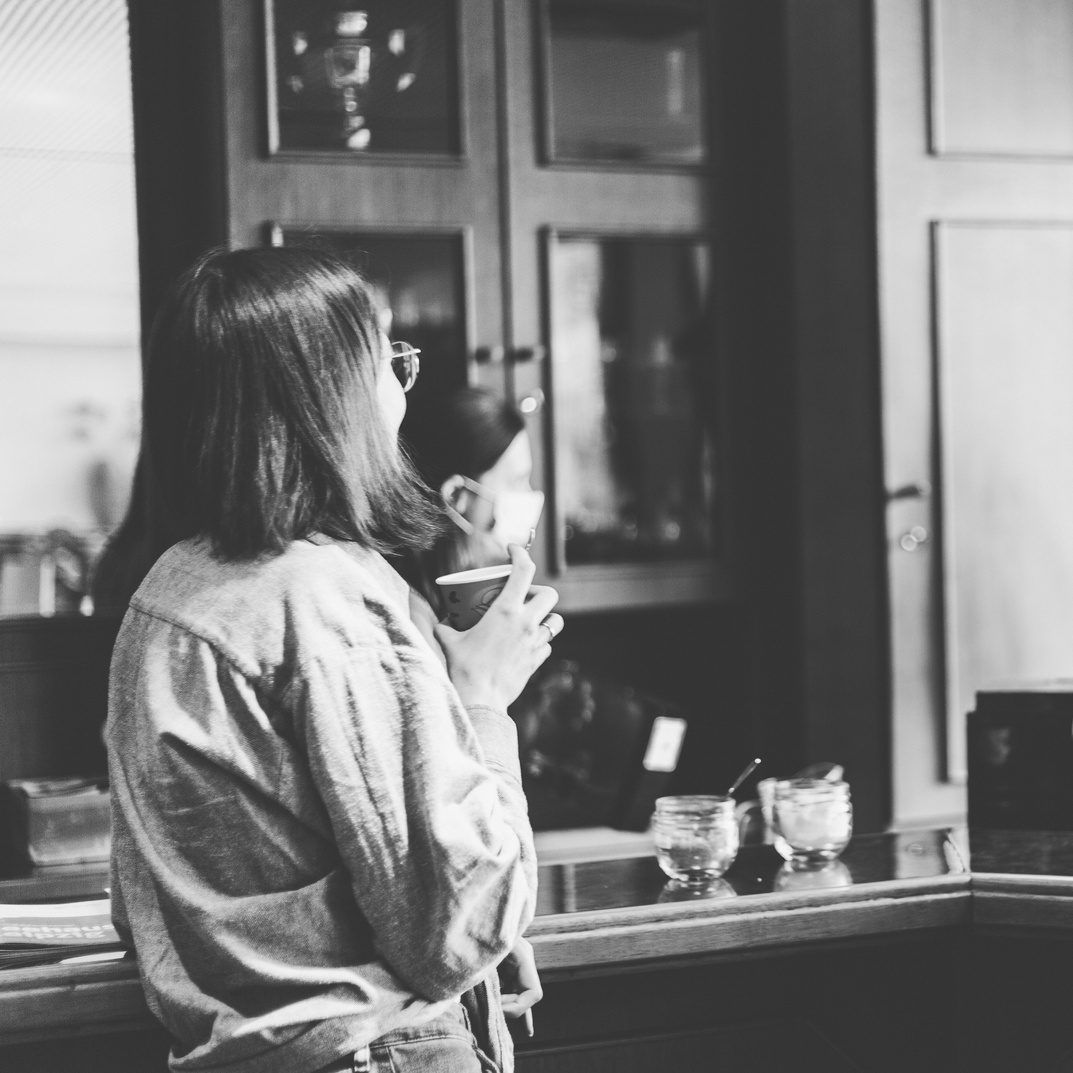 Black-and-white photo of a woman with glasses standing at a counter and drinking from a cup