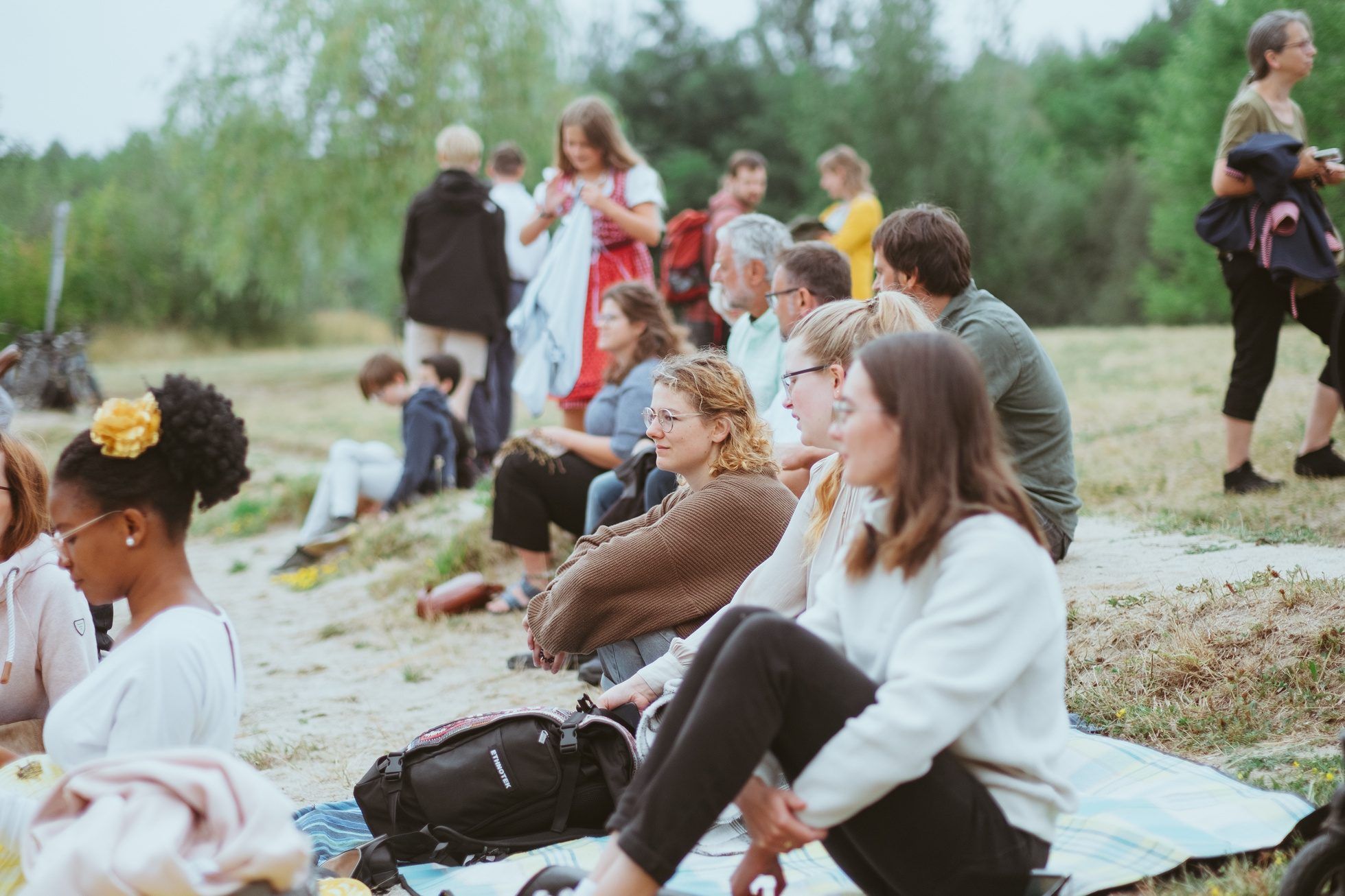 People sit on blankets on a sandy slope outdoors, listening, with others standing in front of trees behind them