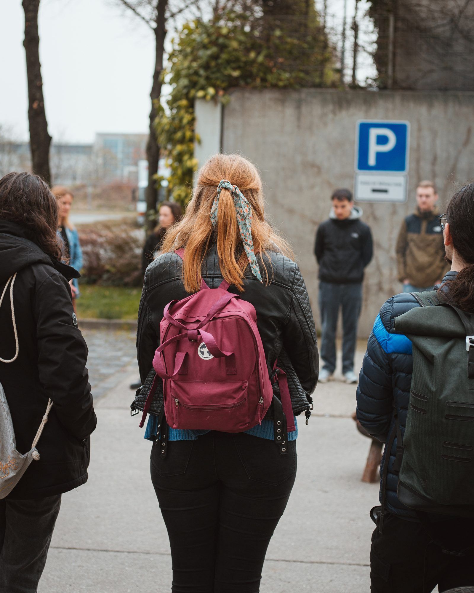 A young woman with strawberry-blonde hair and a red backpack standing in a circle with other people in front of a concrete wall