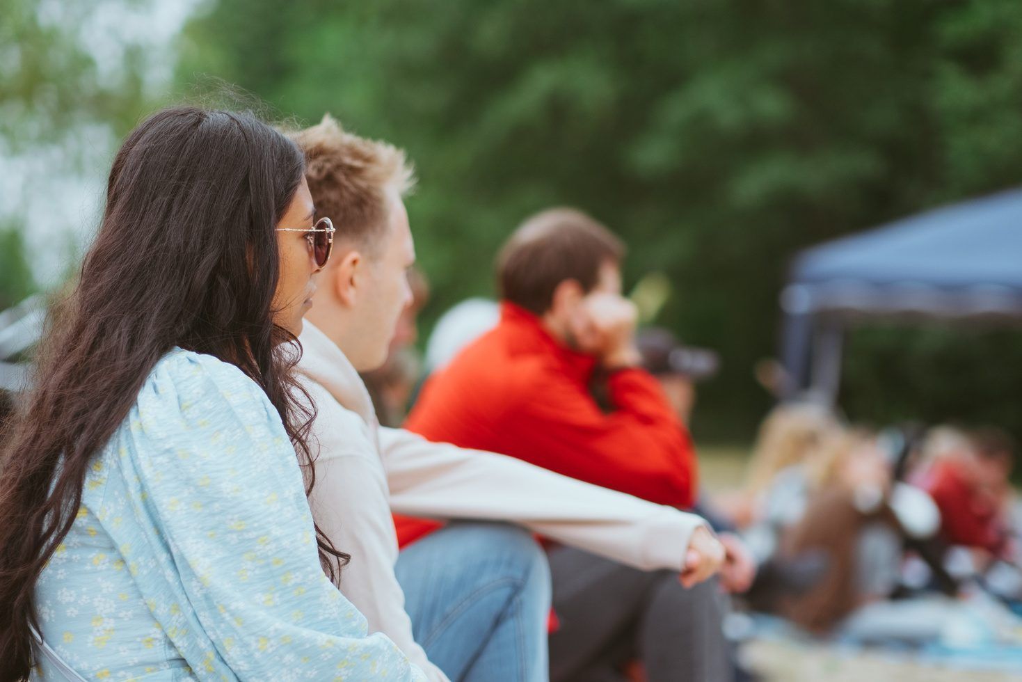 A woman wearing sunglasses and a man sitting outdoors on the grass, with more people and trees in the background