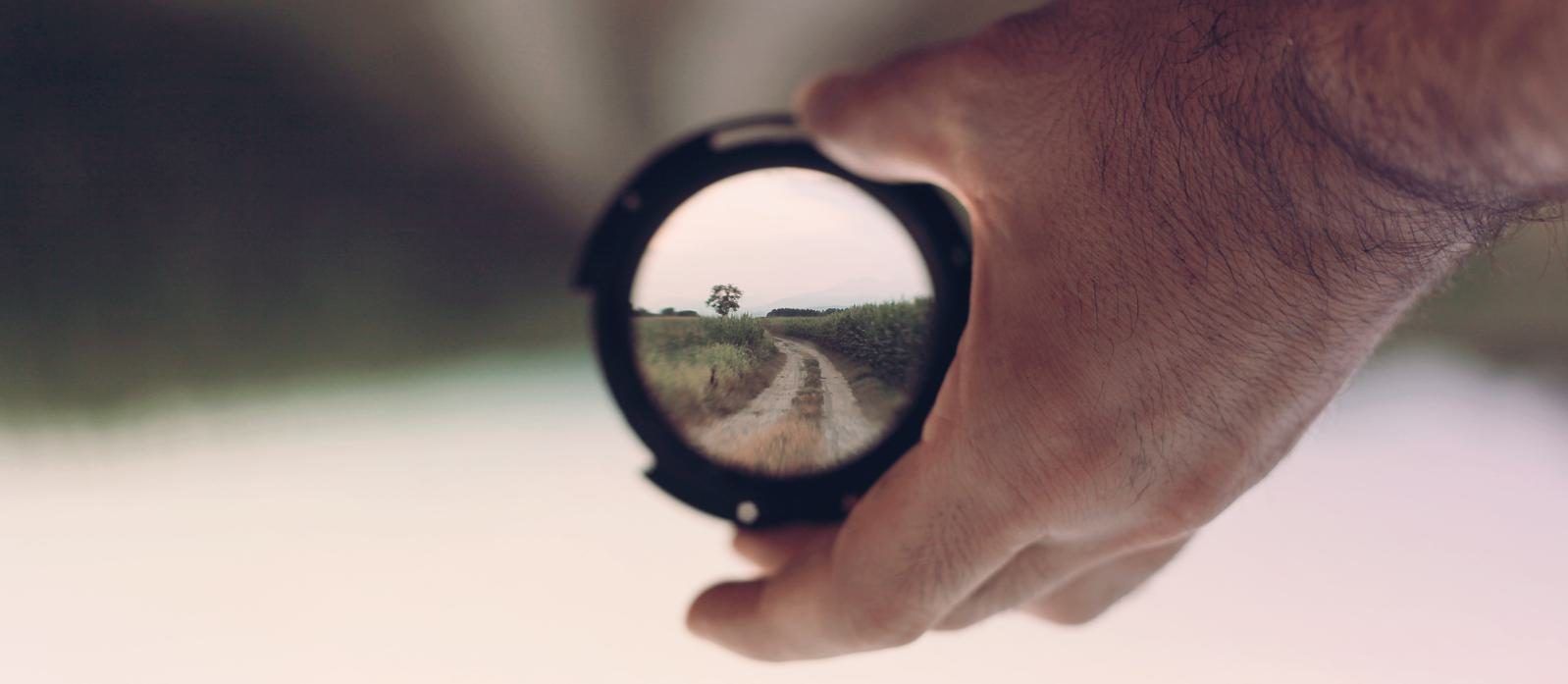 A hand holding a lens through which a dirt path and a lone tree are seen in sharp focus, with the surroundings blurred