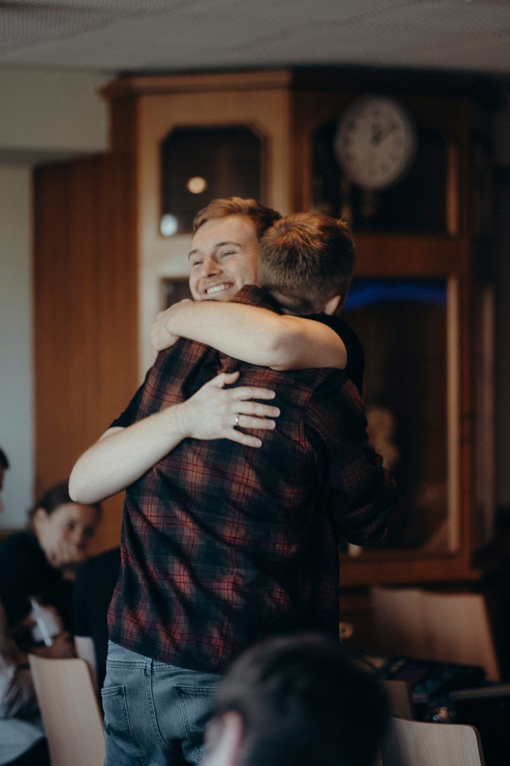 Two men hugging and smiling in a room with a wooden cabinet and a wall clock