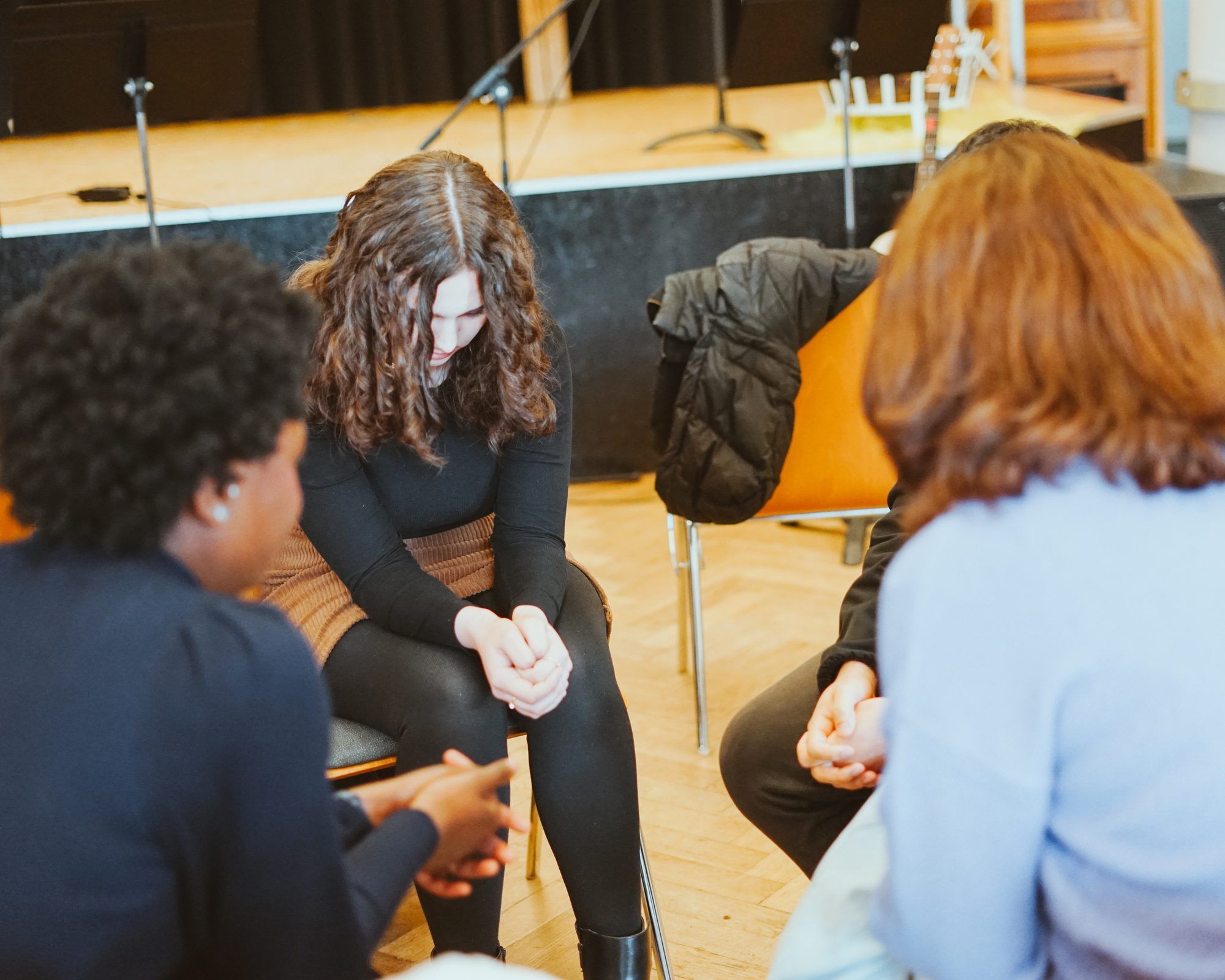 People sitting in a circle on chairs, a young woman with curly hair bowing her head with her hands clasped