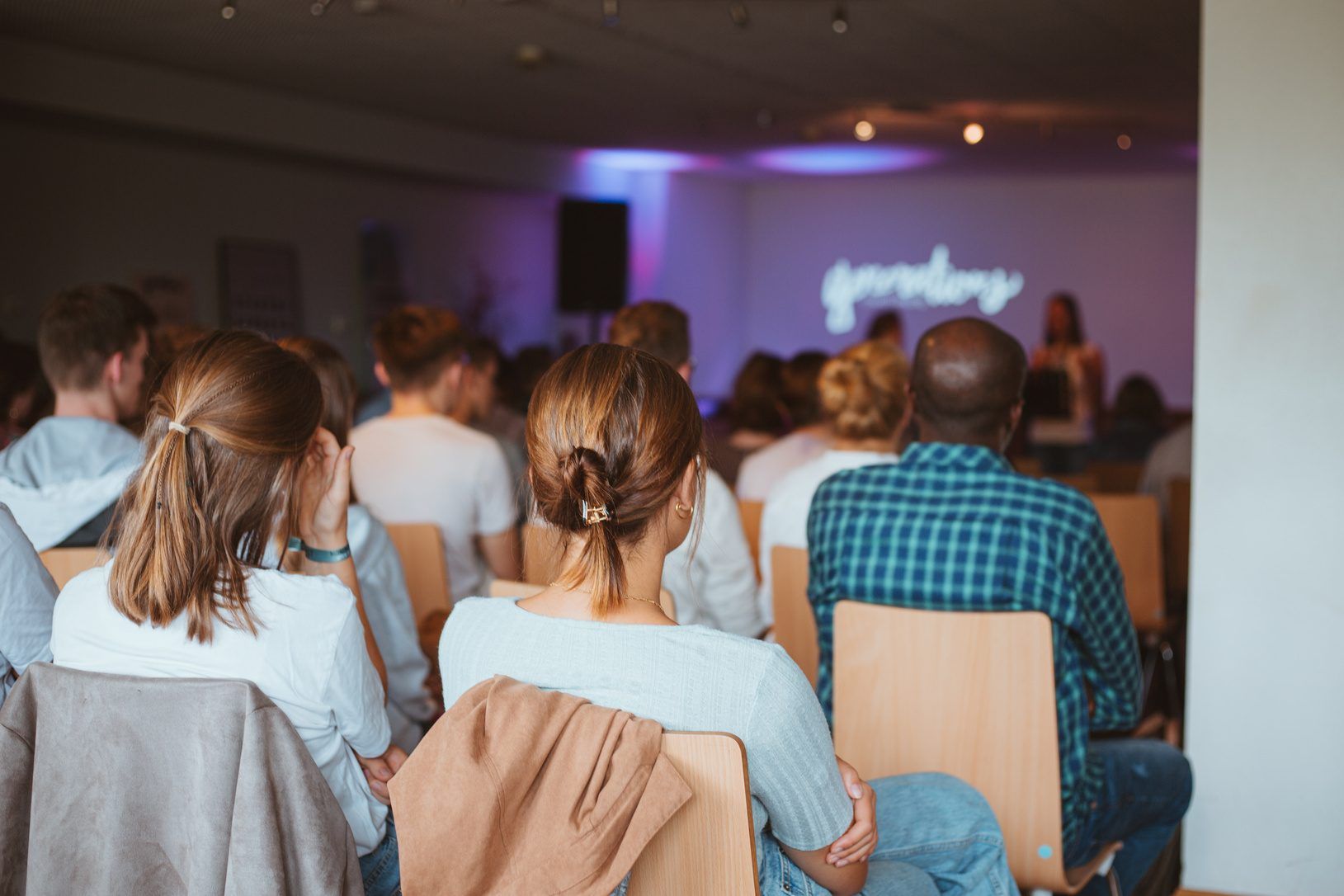 People seen from behind sitting on wooden chairs in a church hall, with a speaker at the front in front of a purple-lit wall