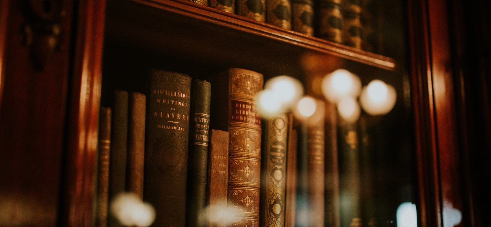 Old leather-bound books on a wooden shelf behind glass, with blurred points of light in front