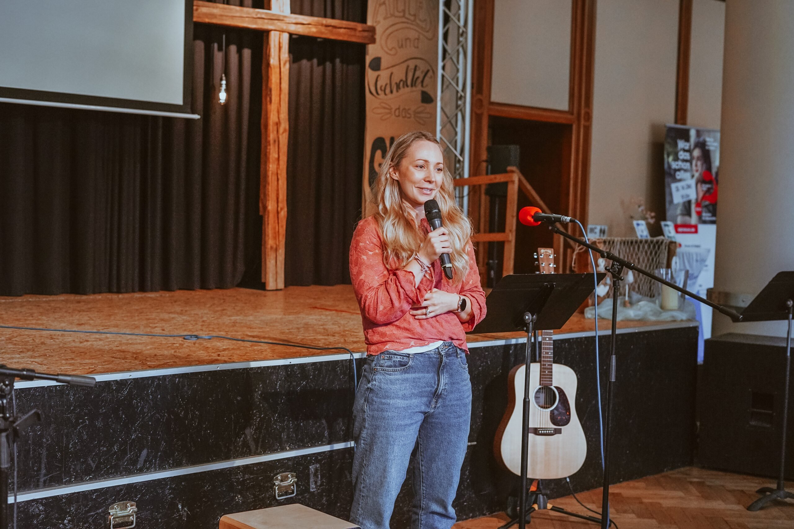 A woman with a microphone speaking on stage, with a guitar beside her