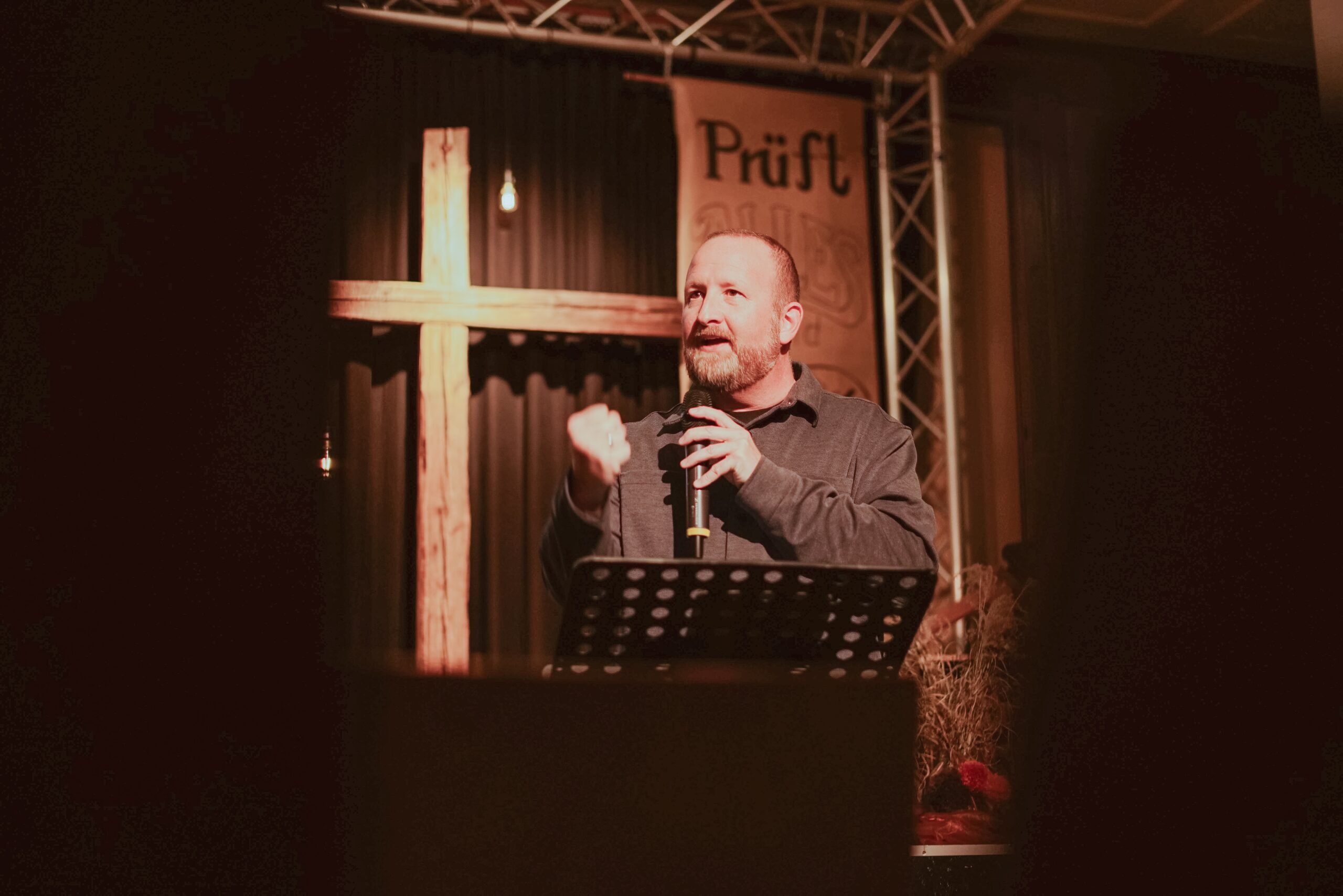 A preacher with a microphone in front of a wooden cross on a dimly lit stage