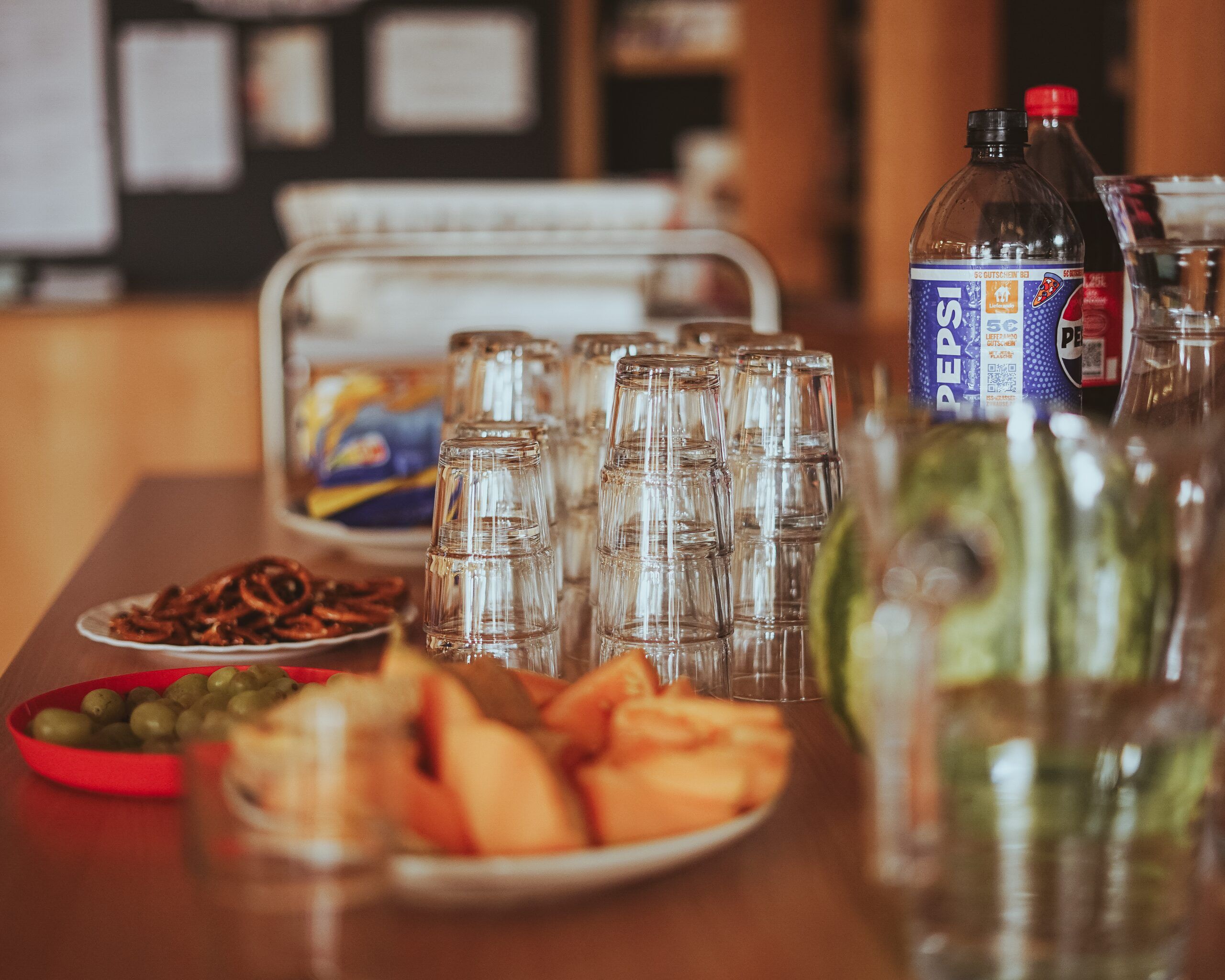 A table with stacked glasses, drinks, pretzels, olives and melon